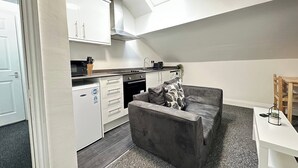 Ett kompakt kök med vita skåp, en inbyggd ugn och en mikrovågsugn. I vardagsrummet finns en grå soffa med en kudde med ett lövmönster, och ett litet träbord med en växt syns.