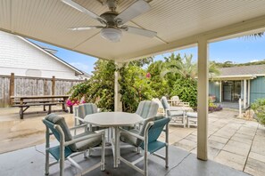 En overdækket terrasse med bord og stole, en loftventilator og udsigt til et hus og en have.