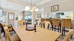A dining area with a wooden table, chairs, and a chandelier. There are framed pictures on the walls and a kitchen area in the background. - 1267 S Fletcher ~ 4 BR House, Walk to the Beach! (Fernandina Beach)