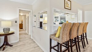 A modern kitchen with white cabinetry, a built-in island, and a dining area with wicker chairs and a yellow cushion. - 1267 S Fletcher ~ 4 BR House, Walk to the Beach! (Fernandina Beach)