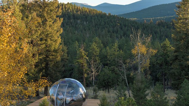 A transparent dome structure on a wooden platform surrounded by a forest with a mountain range in the background. - Hidden Homestead: 5 acres, Luxe Cabin, Sauna, Dome, Gym (Bailey)