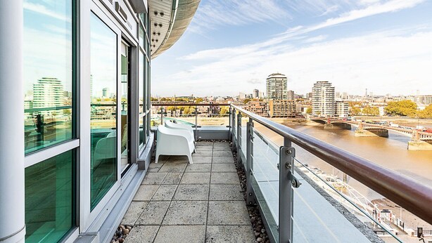 A balcony with a view of a river and cityscape, featuring a modern building with large windows and a white chair. - Riverside Luxury with Iconic Views (London)