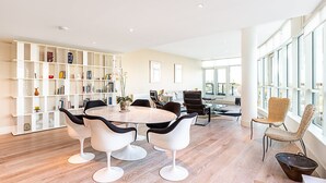 A modern living room with a round table, white chairs, a bookshelf, and large windows. - Riverside Luxury with Iconic Views (London)