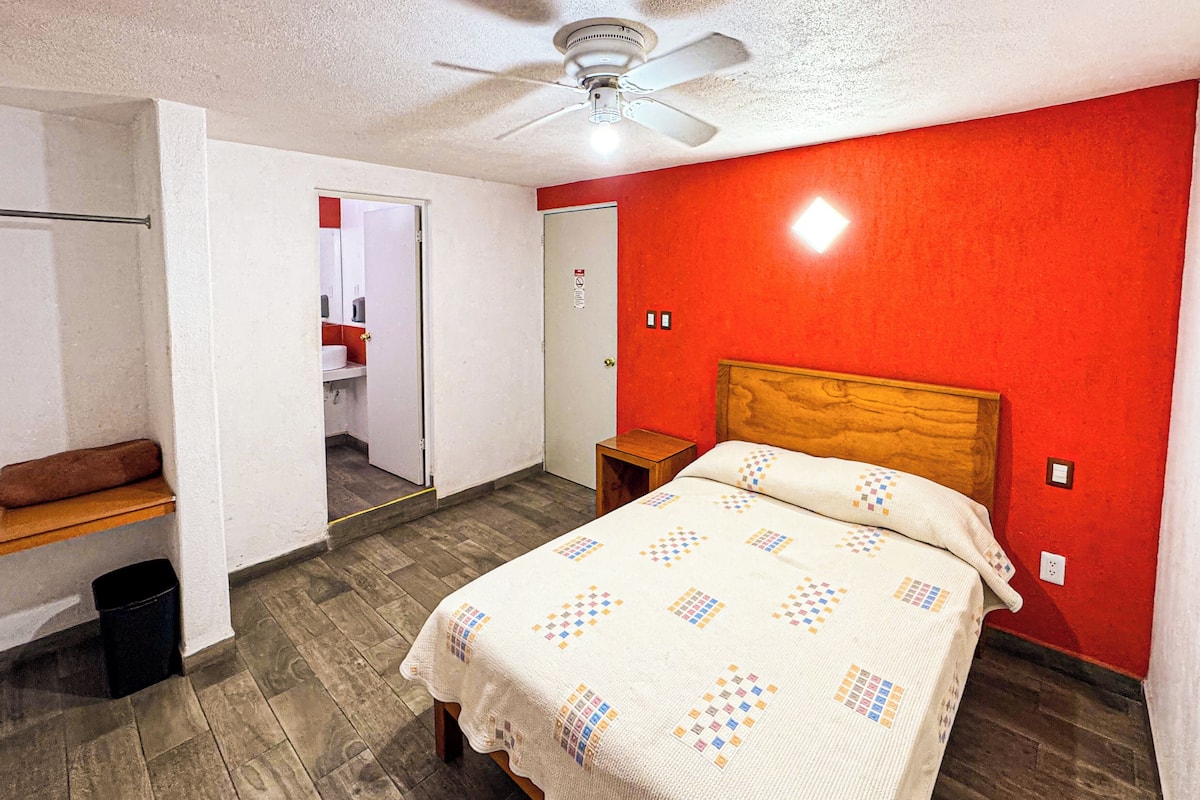 A bedroom with a bed, a wooden headboard, a ceiling fan, a red wall, and a door leading to a bathroom.