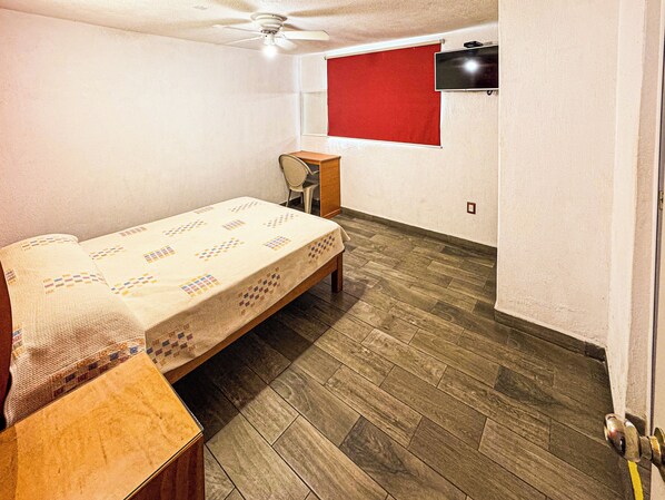 A bedroom with a bed, a desk, a chair, a ceiling fan, a TV mounted on the wall, and a red window blind.