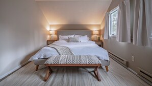 Quarto com cama grande, abajures, um banquinho e janela com cortinas.