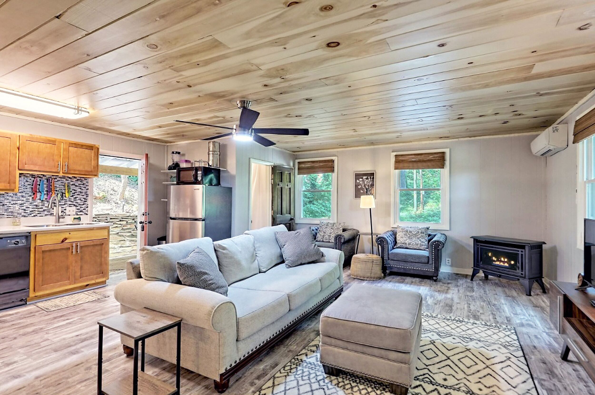A spacious living area with a large sofa, ottoman, and a fireplace. There is a kitchen with wooden cabinets and a stone backsplash.