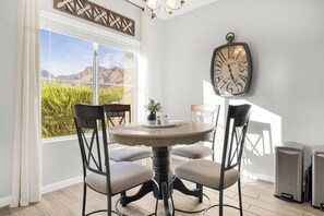 Uma sala de jantar com uma mesa redonda, quatro cadeiras, um relógio na parede e vista para montanhas por uma grande janela.