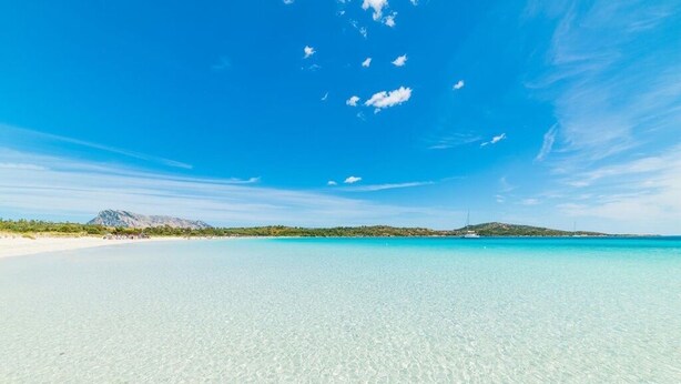 Una spiaggia con acque cristalline turchesi, una montagna all'orizzonte e una piccola isola con un faro.