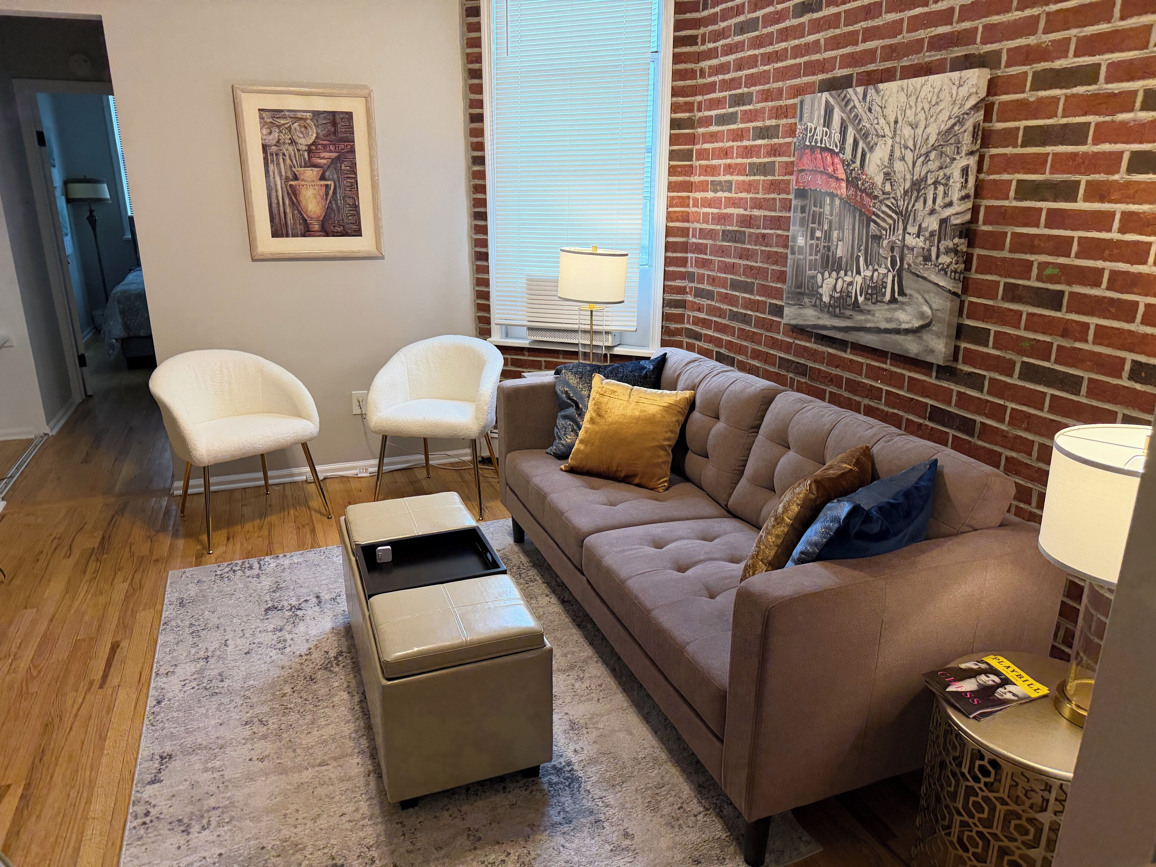 A living room with a beige sofa, two white chairs, a small ottoman, a patterned rug, a lamp, and a painting of Paris on the brick wall.