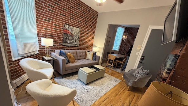A living room with a brick wall, a sofa, two armchairs, a coffee table, and a dining area with a table and chairs.