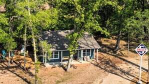 A small, single-story house with a porch, surrounded by trees and a dirt driveway. A 'SKELLY' sign is visible in the foreground. - The Skelly Hideout (Kilgore)