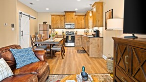 A modern kitchen with wooden cabinets, a dining area with a table and chairs, and a living room with a leather sofa and a television. - The Skelly Hideout (Kilgore)