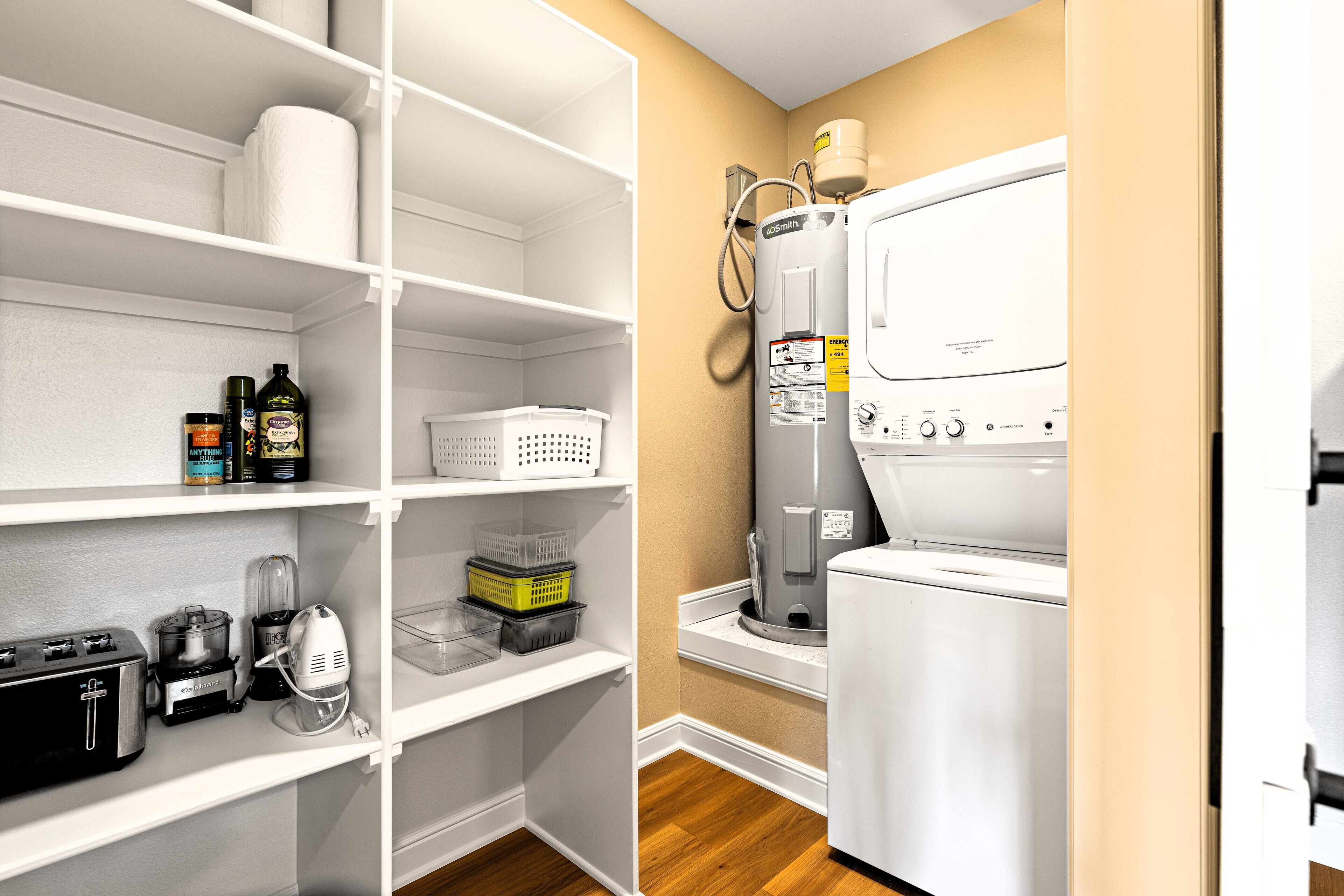 A laundry room with a stacked washer and dryer, a water heater, and various appliances on shelves.