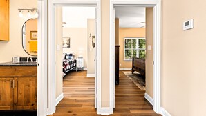 A hallway with wooden flooring, a kitchen area with a sink and cabinets, and a bedroom with a bed and nightstand. - The Skelly Hideout (Kilgore)