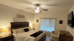 Ein Schlafzimmer mit Bett, Nachttischen, Deckenventilator, Fenster mit Jalousien, einem Holznachtisch und einem an der Wand montierten Fernseher.
