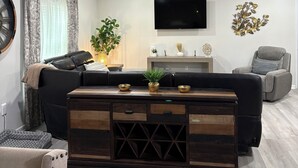 Ein modernes Wohnzimmer mit einem schwarzen Ledersofa, einem Couchtisch aus Holz mit dekorativen Schalen, einem grauen Sessel und einem flachbildfernseher an der Wand.