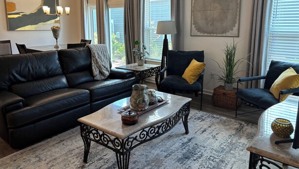 A living room with a black leather sofa, a coffee table, and two armchairs. There are large windows with curtains, a painting on the wall, and a chandelier. - 3/2 Villa in most desired Richmond neighborhood in the Villages. Jan-Apr rental (The Villages)