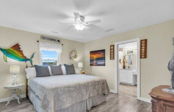 A bedroom with a bed, a ceiling fan, a window with a view, a vanity with a mirror, and a wall decoration. - Gone Coastal- Tranquil 3 Bedroom House with access to the White Oak River (Cedar Point)