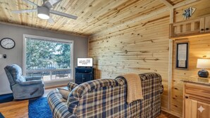 L’intérieur d’un chalet accueillant, avec un canapé à carreaux, un fauteuil inclinable, un téléviseur et une grande fenêtre donnant sur la forêt.