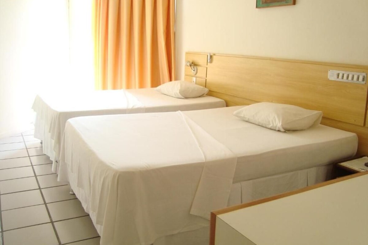 A hotel room with two single beds, a wooden headboard, a white bedside table, and a window with orange curtains.