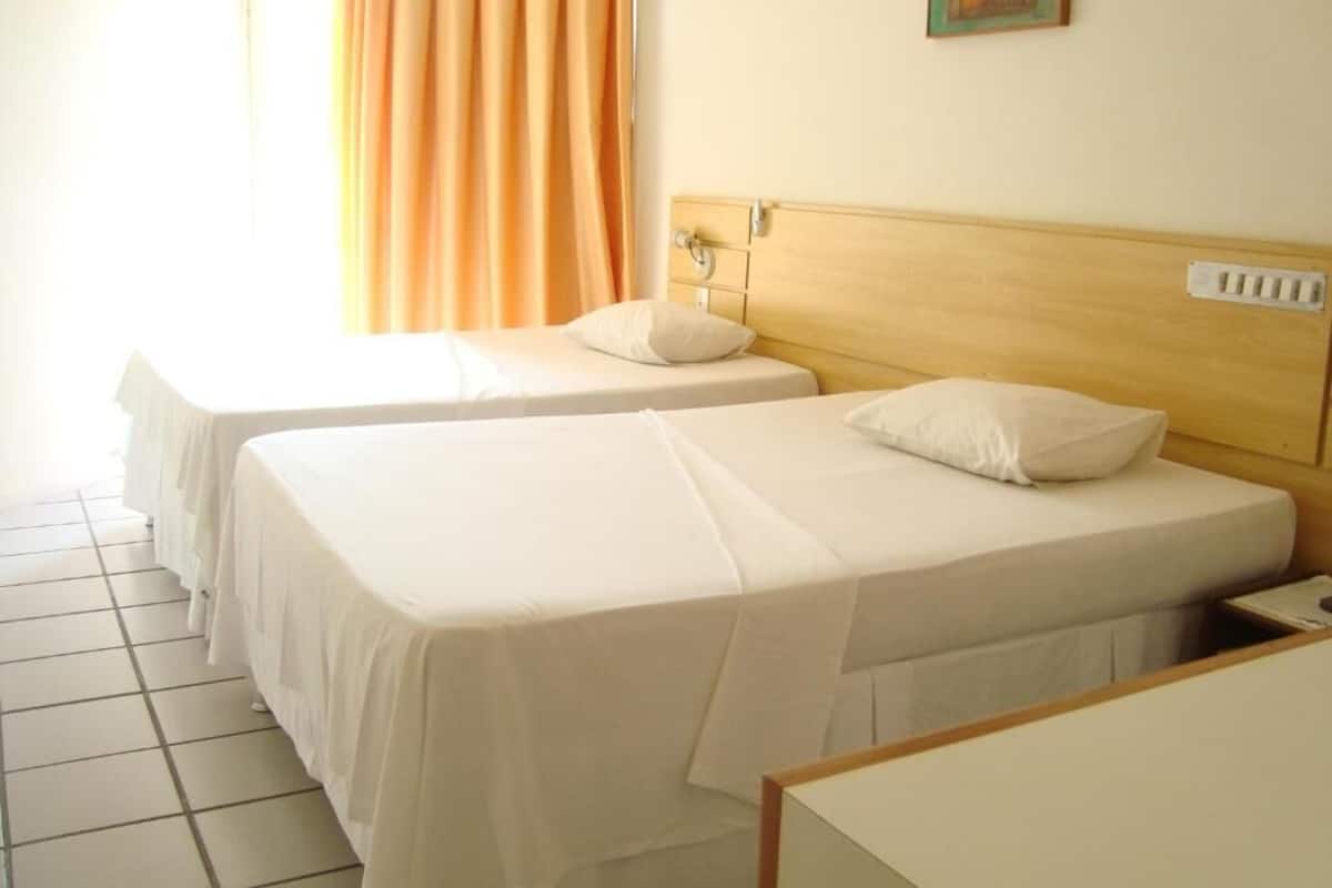 A hotel room with two single beds, a wooden headboard, a white bedside table, and a window with orange curtains.