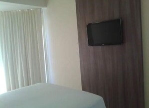 Una habitación de hotel con una cama, un televisor montado en un panel de madera y una ventana con cortinas.
