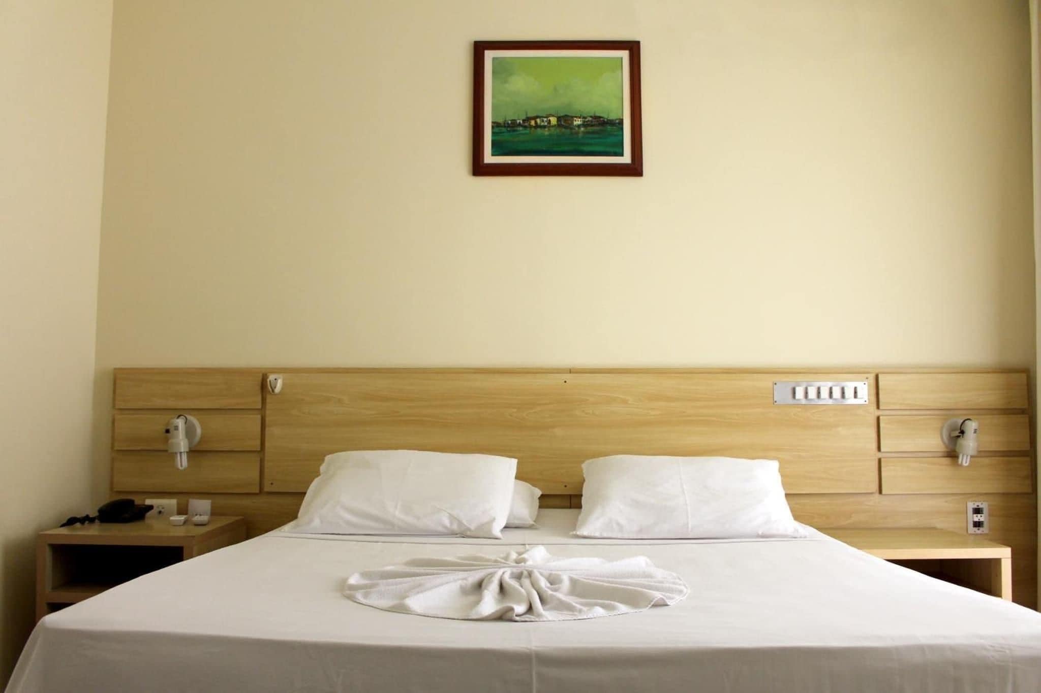 A hotel room with a wooden headboard, white bedding, a neatly made bed, a bedside table with a lamp, and a painting on the wall.