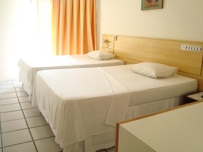 Una habitación de hotel con dos camas individuales, un cabecero de madera, una mesita de noche pequeña y una ventana con cortinas.