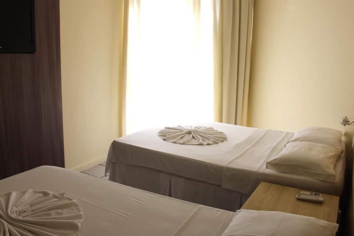 A hotel room with two beds, a wooden nightstand, a remote control, and a window with curtains.