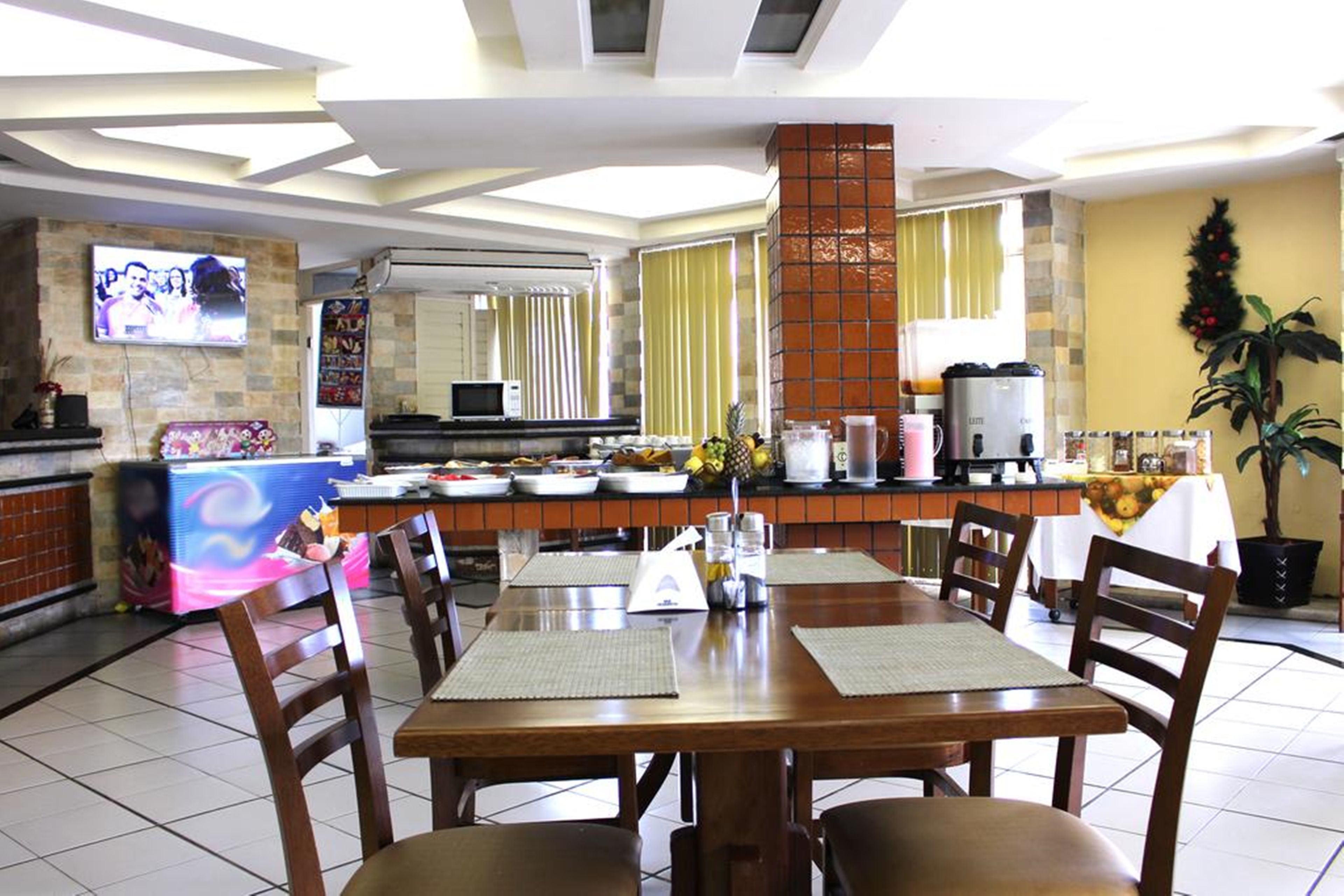 Um restaurante com área de jantar, mesa e cadeiras de madeira grande, cozinha ao fundo e uma TV na parede.