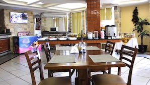 Un restaurant amb una zona de dinar que destaca per una gran taula i cadires de fusta, una cuina al fons i un televisor penjat a la paret.