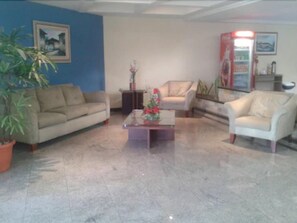 A living room with a beige sofa, a white armchair, a coffee table with a flower arrangement, and a potted plant. - Rede Andrade Aracaju (Aracaju)