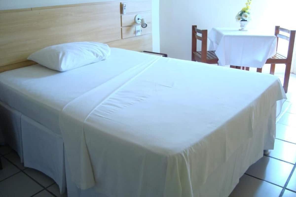 A neatly made bed with white bedding, a wooden headboard, and a small table with a white tablecloth and a vase of flowers in the background.