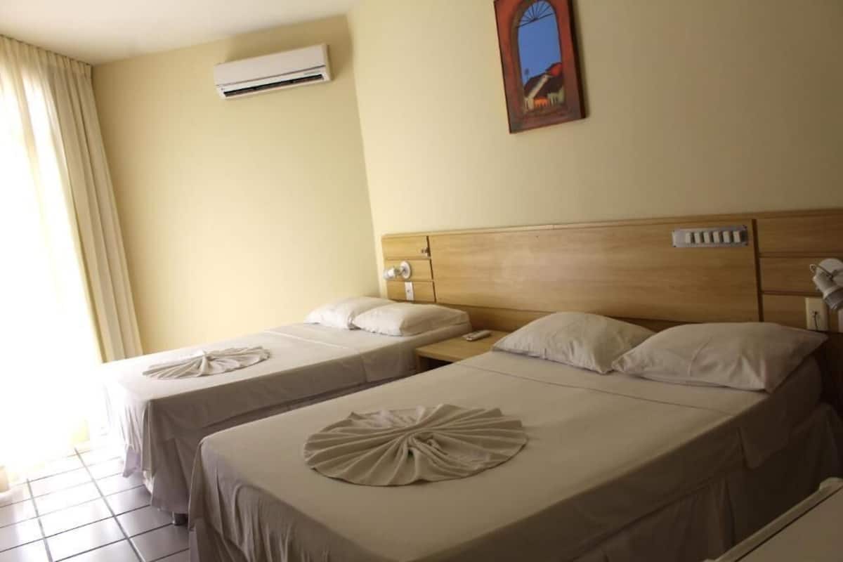 A hotel room with two beds, each with a neatly made bedspread and a decorative pillow. There is an air conditioning unit on the wall and a painting above the beds.