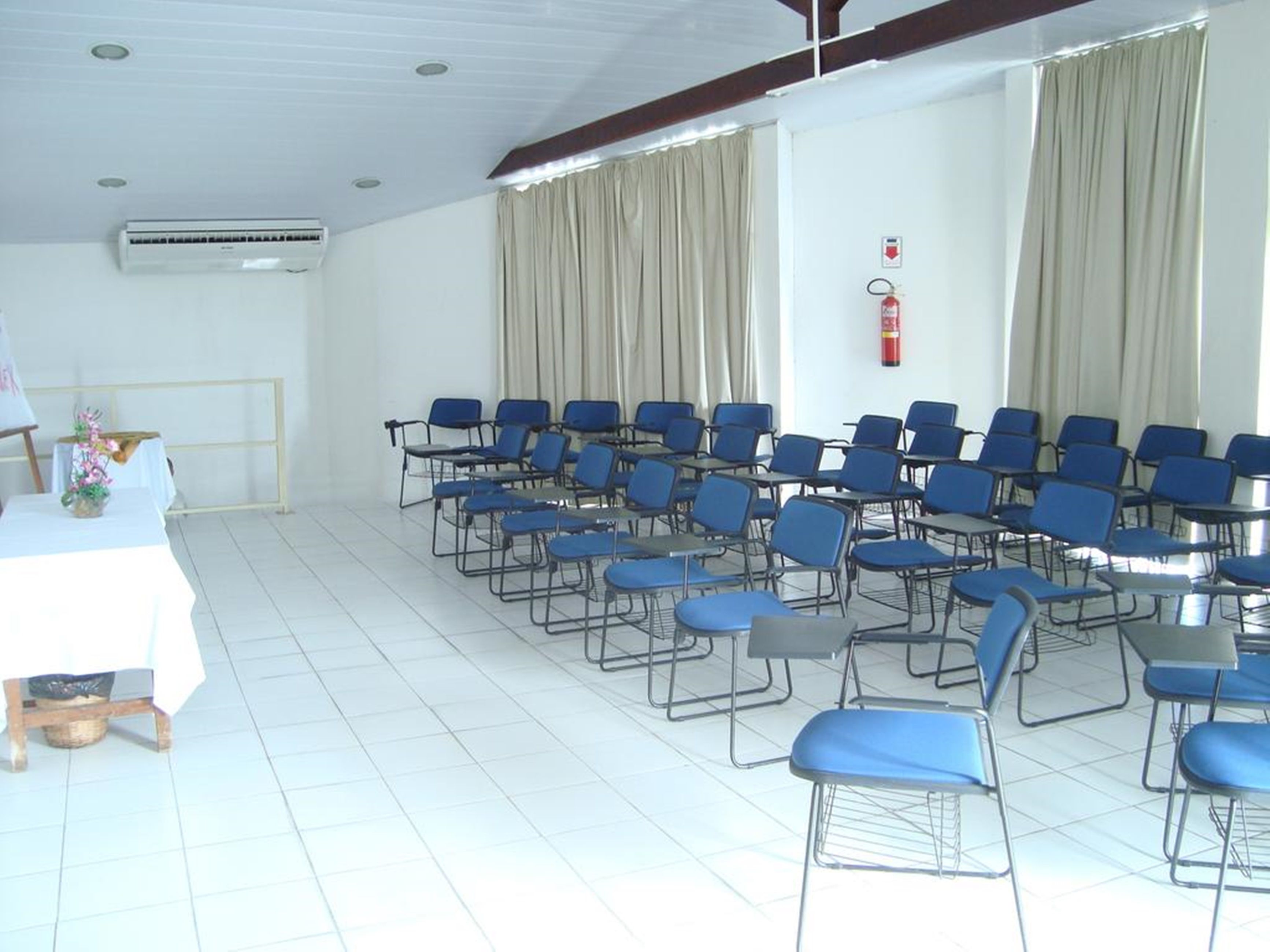 Uma sala de conferências com fileiras de cadeiras azuis, uma mesa branca com um vaso de flores e um extintor na parede.