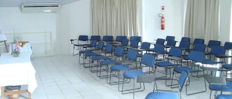 Un salón de conferencias con filas de sillas azules, una mesa blanca con un jarrón de flores y un extintor en la pared.