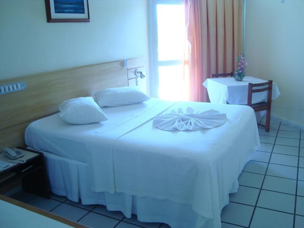 A hotel room with a bed, a bedside table with a phone, a small table with a vase, and a window with curtains.
