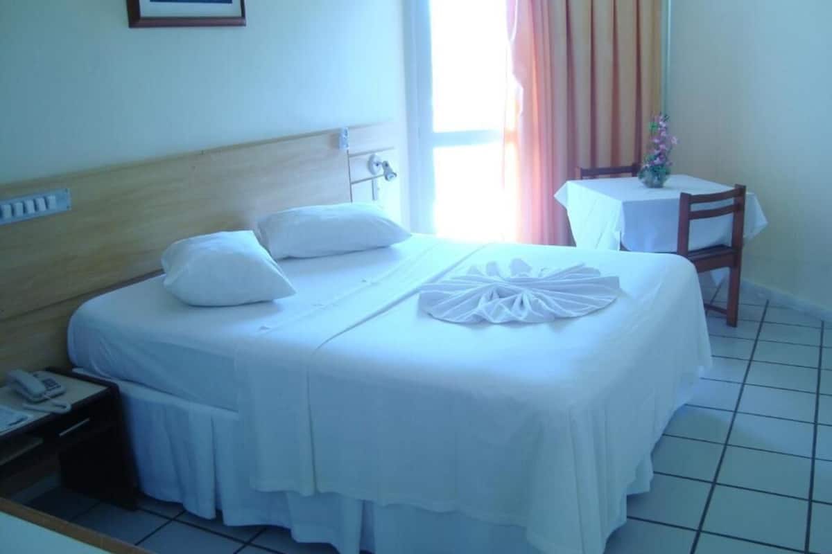 A hotel room with a bed, a bedside table with a phone, a small table with a vase of flowers, and a window with curtains.