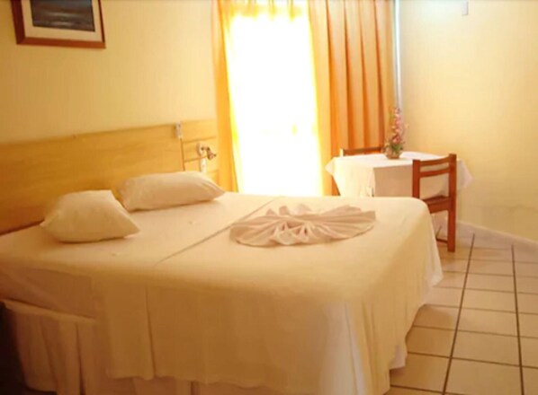 A neatly made bed with pillows and a folded towel, a small table with a vase, and a chair in a room with a tiled floor and a window with curtains.
