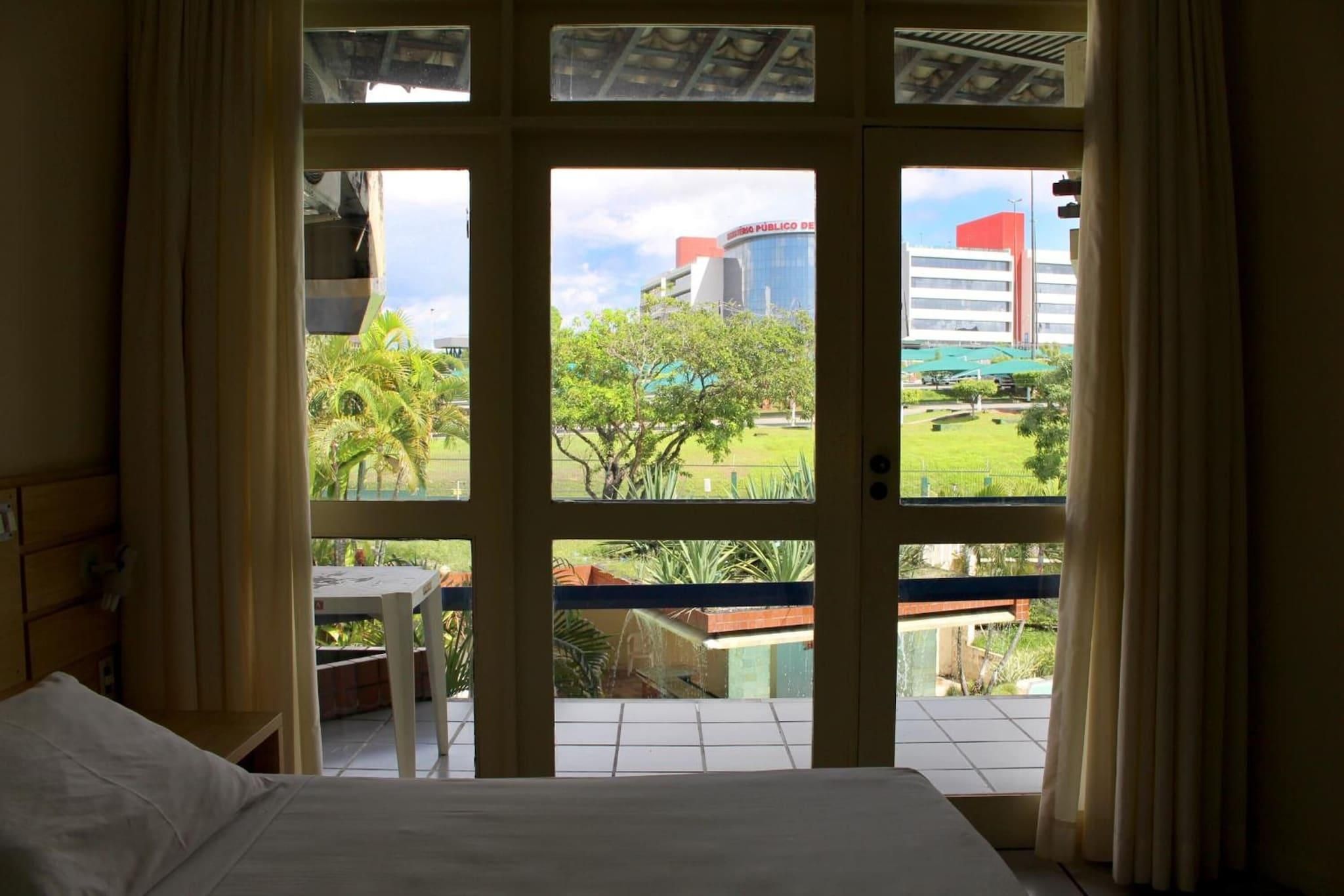 Quarto de hotel com uma cama, uma mesinha e vista para um edifício e vegetação lá fora.