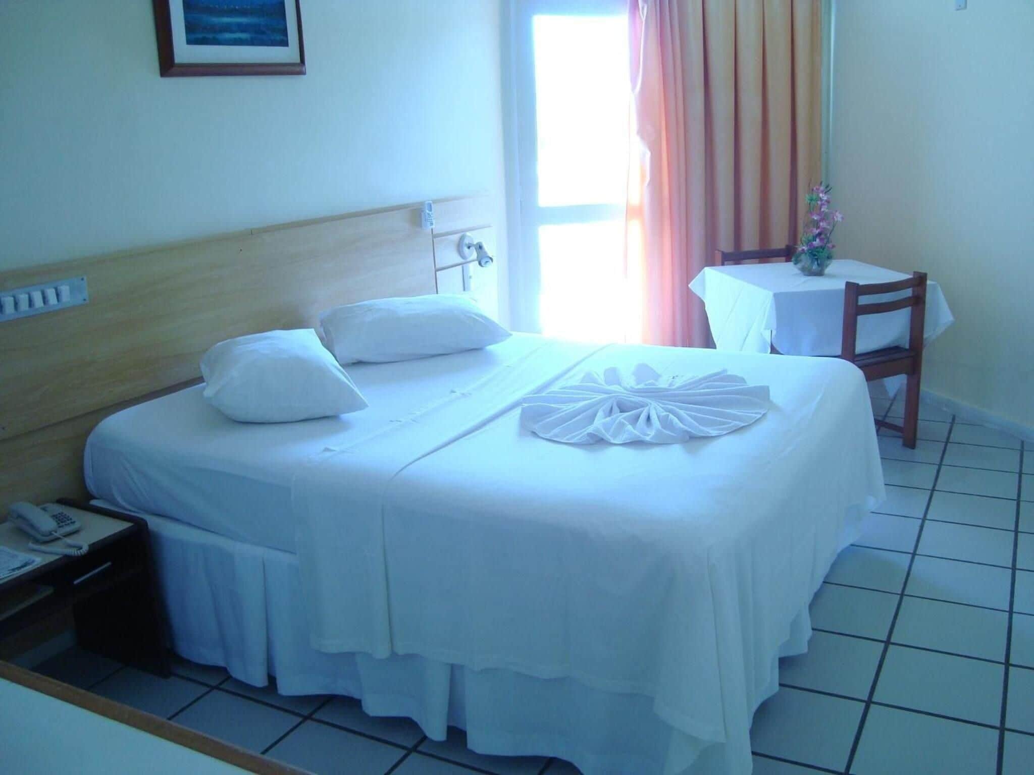 Quarto de hotel com uma cama, mesa de cabeceira com telefone, pequena mesa com um vaso e janela com cortinas.