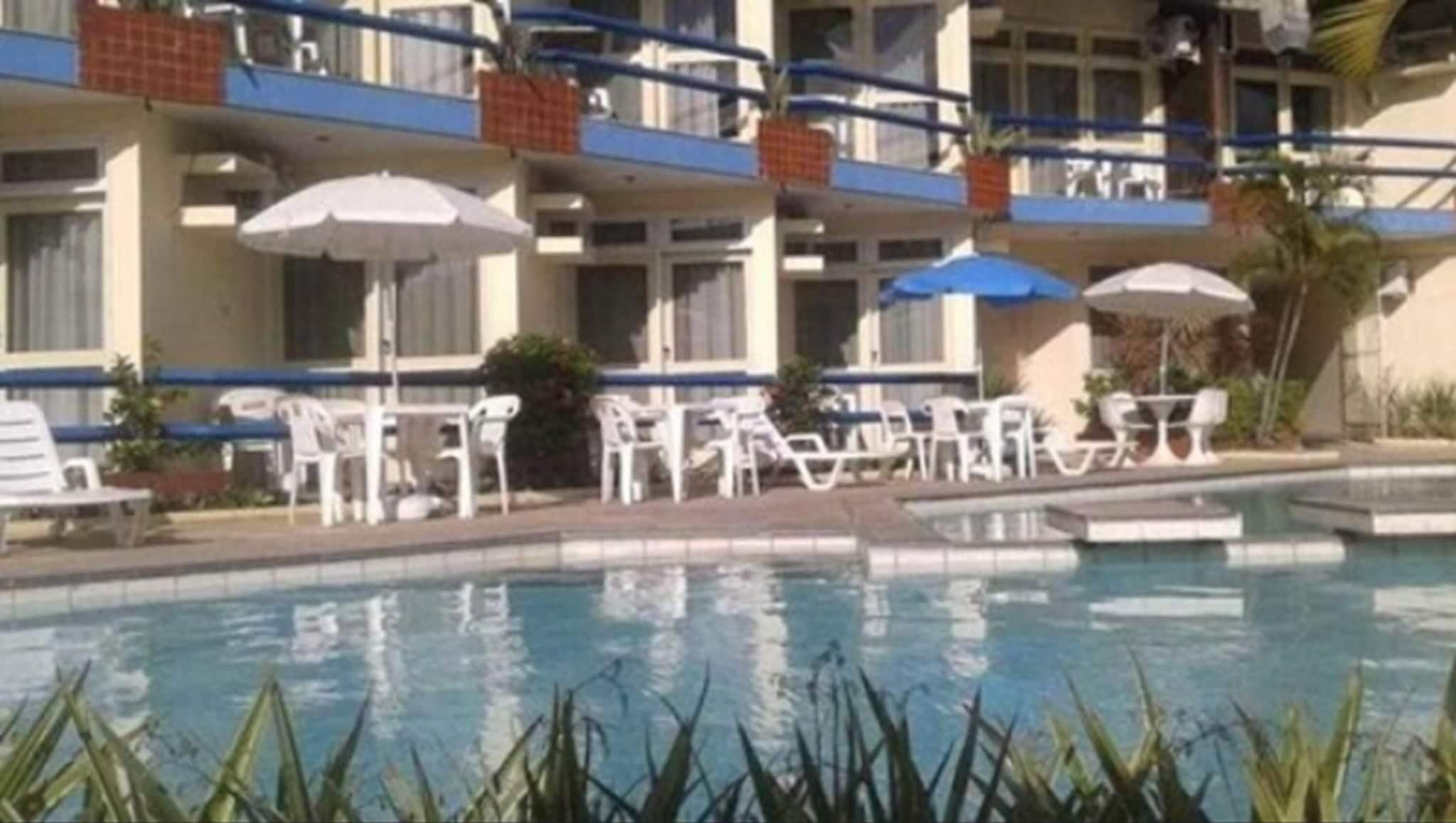 Una piscina con sillas de descanso y sombrillas, rodeada por un edificio con balcones y ventanas.