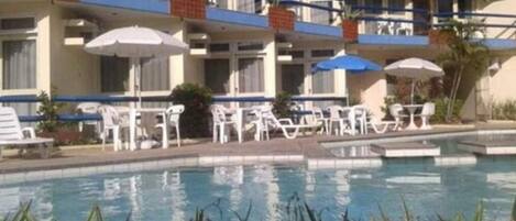 Una piscina con sillas de descanso y sombrillas, rodeada por un edificio con balcones y ventanas.