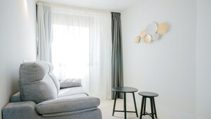 A modern living room with a grey sofa, two black stools, a large window with sheer curtains, and a wall-mounted decorative piece. - Hotel Cullera Coast by Mij (Cullera)