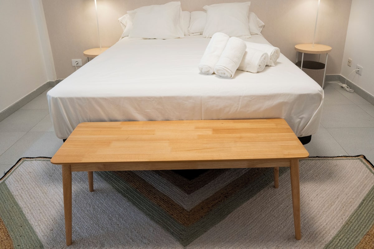 A neatly made bed with white linens and rolled towels, a wooden bench in front, and two bedside lamps on either side.