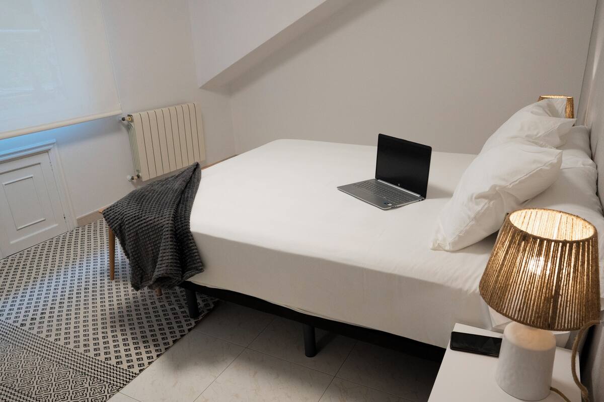 A neatly made bed with a laptop, a lamp, and a patterned rug.