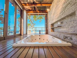 A hot tub in a room with a wooden floor, stone walls, and large windows overlooking a balcony with a view of the sea. - Hotel Nacional Inn Ubatuba Praia Das Toninhas (Ubatuba)