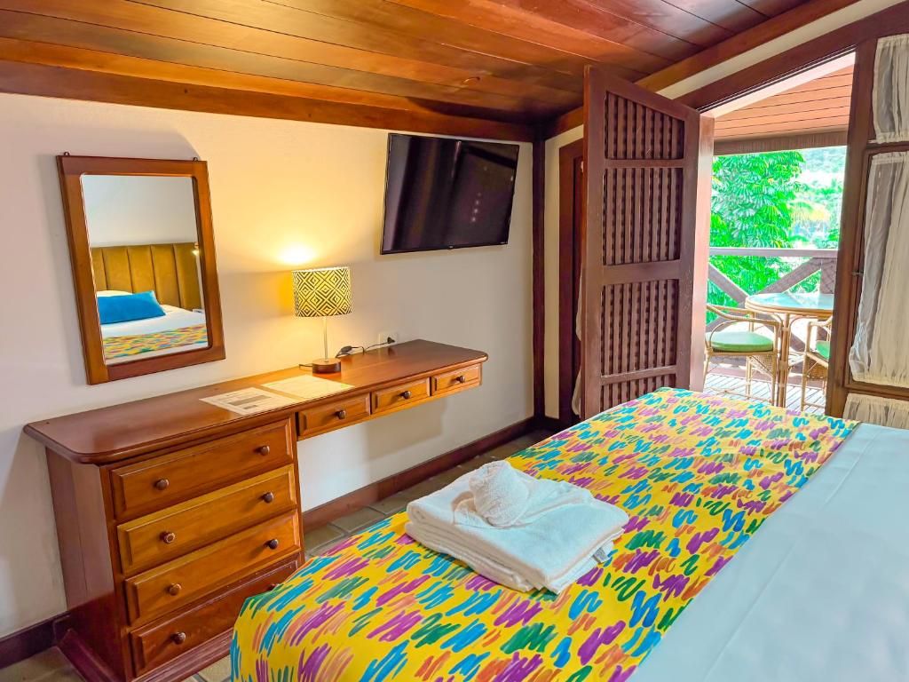 A hotel room with a bed, a wooden dresser, a television, and a window with a view of greenery.