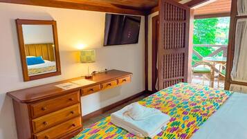 A hotel room with a bed, a wooden dresser, a television, and a window with a view of greenery.